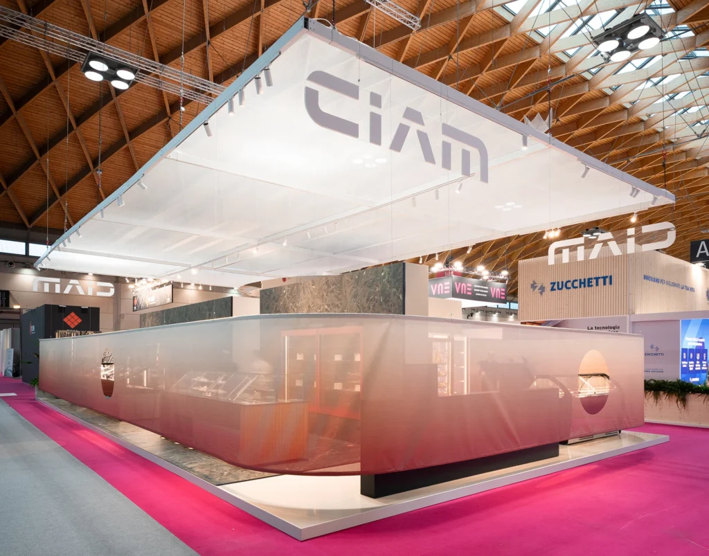 A modern exhibition booth with frosted glass walls and a suspended white canopy displaying the logo CIAM. The booth is set up in a large, well-lit convention center with pink carpeting.