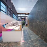 A modern, empty glass display case stands on a blue-gray marble floor next to a high marble wall in a brightly lit indoor space with large windows and a pink carpet on the left side.