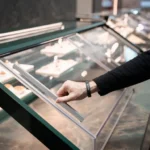 A person lifts a clear glass or plastic cover on a display case filled with plated food items, possibly in a cafeteria or buffet setting. The person is wearing a black long-sleeve shirt and a watch.