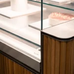A modern glass display case with wooden panels showcases assorted pastries and desserts on white trays, including tarts and cakes, in a well-lit bakery or café setting.