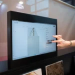 A person’s hand selects a color option on a touchscreen displaying material samples, with stone slabs displayed below the screen in a modern showroom.