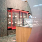 A man stands in a modern bakery with marble walls and floors, looking at mostly empty red-lit display refrigerators. A glass case with a few pastries is in the foreground.