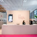A modern exhibition booth with a large counter, two people talking, and a wall displaying “Rethinking Intelligence.” The space is minimalistic, with plants, bright lighting, and digital displays on the side wall.