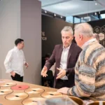 Two men stand at a circular interactive display table, engaged in conversation. One gestures with his hands while the other listens. Other people are visible in the background of the modern, well-lit room.
