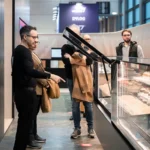 Several people stand in front of a refrigerated display case, looking at items inside. One man gestures toward the case while others watch. The setting appears to be modern and well-lit, possibly a market or exhibition.
