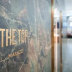 Close-up of a textured wall with the words Thanks to THE TOP MARAZZI in gold letters. The background is blurry, showing a modern, well-lit interior corridor.