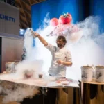 A chef demonstrates a culinary technique with liquid nitrogen, creating clouds of vapor around a table with tins and a book, while a large screen behind displays a dessert topped with strawberries.