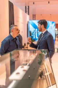 Two men are having a conversation indoors next to a glass display case. One man wears a suit and tie, the other wears a dark button-up shirt. The setting appears to be modern and professional.