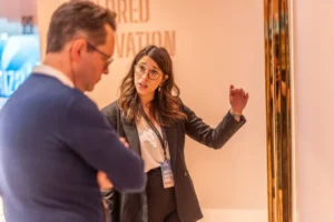 A woman in business attire gestures while speaking to a man in a blue sweater, who listens with his arms crossed. They appear to be having a discussion in a professional setting.