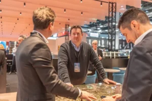 Three men in business attire have a discussion around a marble table at a busy, modern indoor event or conference. One man stands facing the camera while the other two face away. Other attendees are visible in the background.