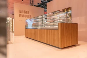A modern glass display case with wooden paneling showcases pastries in a sleek, minimalist store; the wall behind reads “TAILORED INNOVATION.”.