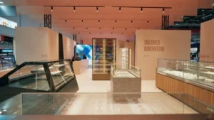 Modern exhibition booth with glass display cases and a minimalist design. The space features large beige walls, bright lighting, and the words Tailored Innovation printed on one wall.
