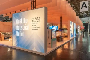 A modern exhibition booth with a large sign reading Meet the Industrial Atelier and CIAM INDUSTRIE, surrounded by display cases and seating, set in a brightly lit convention hall.